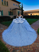 A woman stands outdoors on a brick path in front of a villa, wearing an oversized light blue ball gown with a dramatic train and voluminous layered skirt. / This gown features an off-the-shoulder bodice with shimmering embroidery and a full, layered ball-gown skirt. Ruffled tiers and floral accents add volume, while a long, flowing train completes the elegant look.