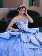 Woman in a blue formal gown with a tiara and jewelry, standing outdoors. / This gown features an off-the-shoulder bodice with shimmering embroidery and a full, layered ball-gown skirt. Ruffled tiers and floral accents add volume, while a long, flowing train completes the elegant look.