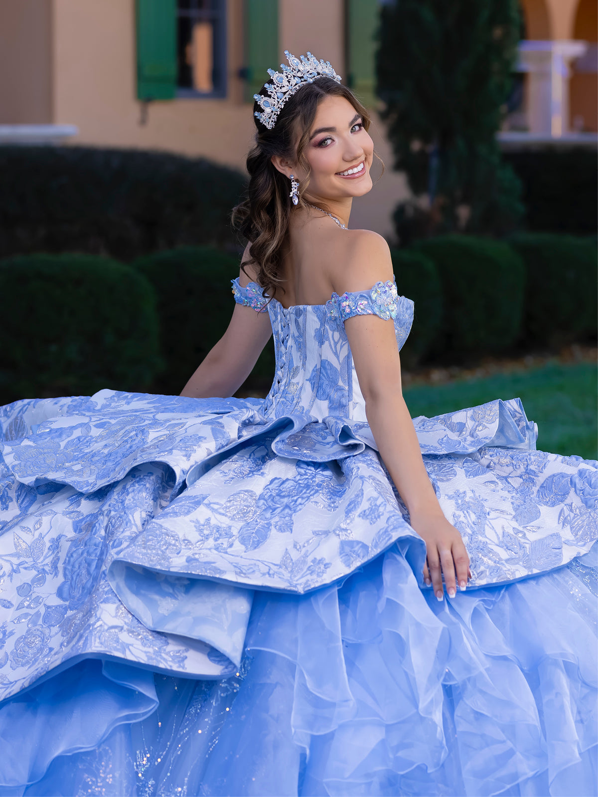 Woman in a blue floral off-shoulder gown with a tiara, sitting outdoors. / This gown features an off-the-shoulder bodice with shimmering embroidery and a full, layered ball-gown skirt. Ruffled tiers and floral accents add volume, while a long, flowing train completes the elegant look.