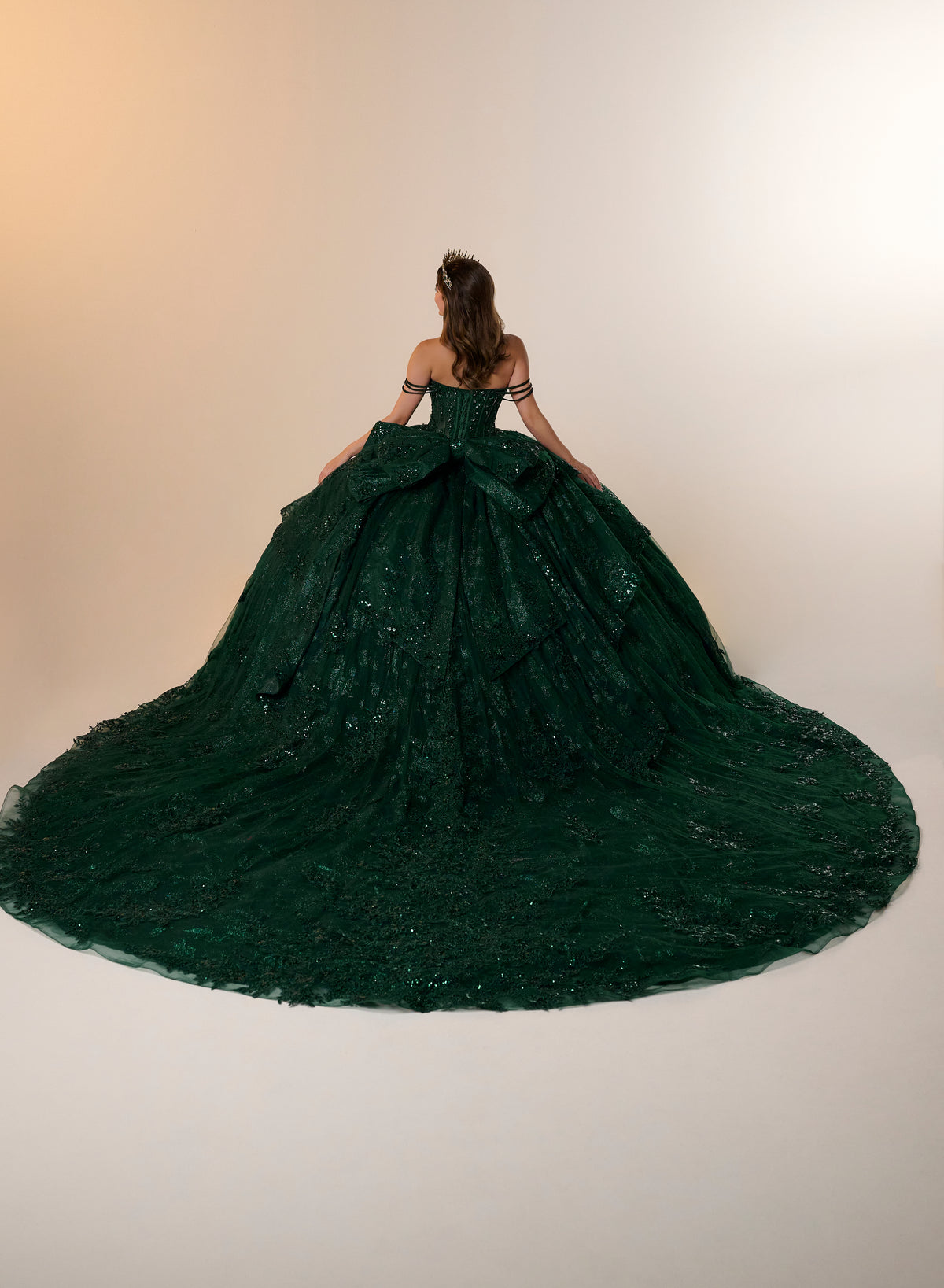 A woman viewed from behind wearing an off-the-shoulder emerald green ball gown with a massive layered tulle train against a plain light beige studio background. / Glamorous ball gown featuring a sparkling embellished corset bodice, dramatic layered overskirt details, a voluminous skirt, and an elegant sweeping train.