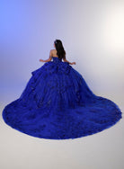 A woman wearing a voluminous royal blue ball gown with intricate beadwork and a long train, photographed from behind against a plain studio backdrop. / Glamorous ball gown featuring a sparkling embellished corset bodice, dramatic layered overskirt details, a voluminous skirt, and an elegant sweeping train.