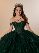 A smiling young woman wearing a sparkling off-the-shoulder emerald green quinceañera dress with sequins, a matching green gemstone tiara, and a green gemstone necklace, posed against a solid beige studio background. / Glamorous ball gown featuring a sparkling embellished corset bodice, dramatic layered overskirt details, a voluminous skirt, and an elegant sweeping train.