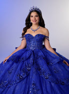 A young woman wearing an elaborate royal blue ball gown with beaded detailing, a matching tiara, and a blue beaded necklace, posed against a soft blue-to-gray gradient studio background. / Glamorous ball gown featuring a sparkling embellished corset bodice, dramatic layered overskirt details, a voluminous skirt, and an elegant sweeping train.