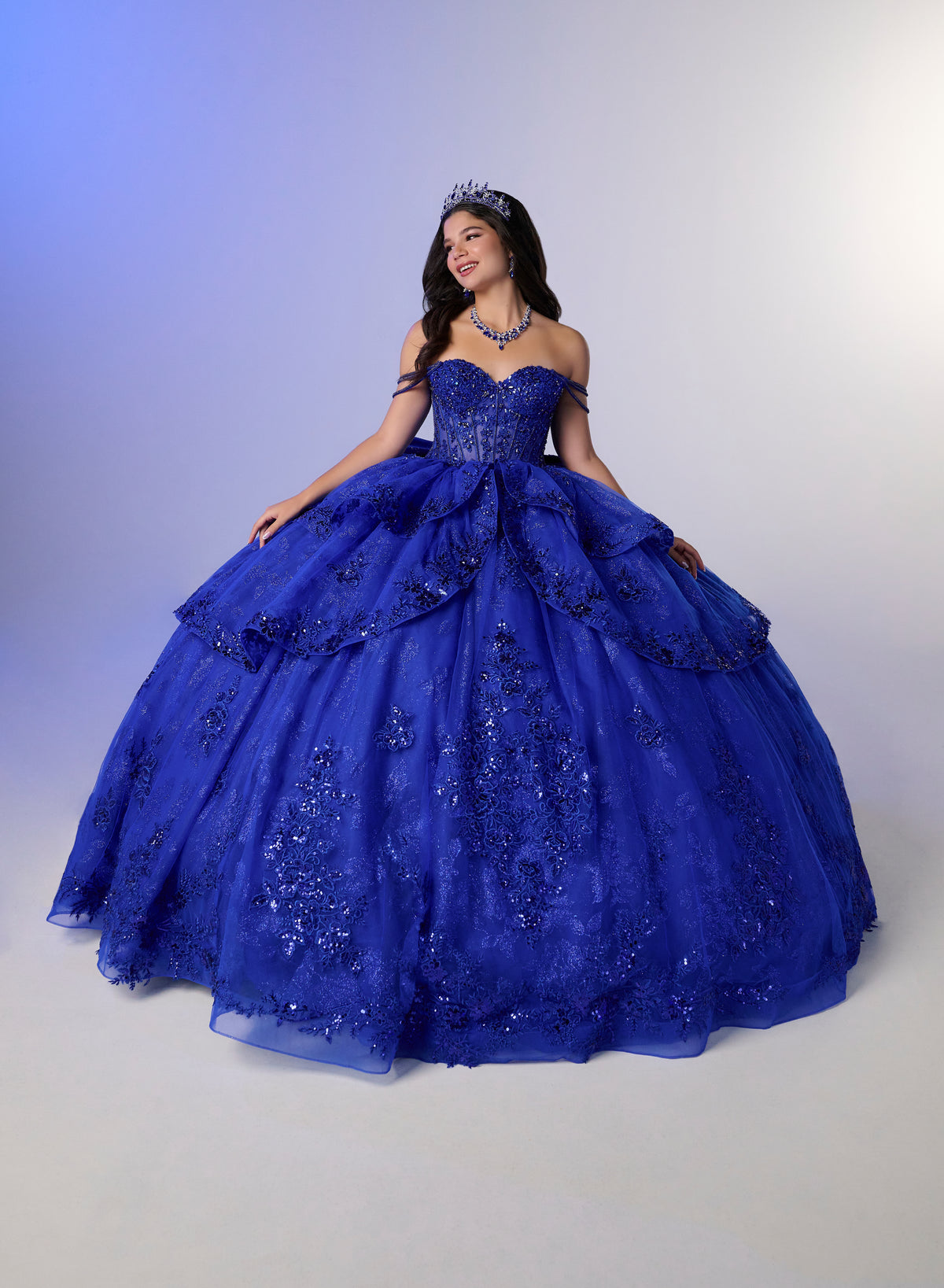 A young woman wearing a voluminous royal blue quinceañera ball gown with floral lace applique, a sweetheart neckline, and a tiara, posing against a studio backdrop with a blue-to-white gradient. / Glamorous ball gown featuring a sparkling embellished corset bodice, dramatic layered overskirt details, a voluminous skirt, and an elegant sweeping train.