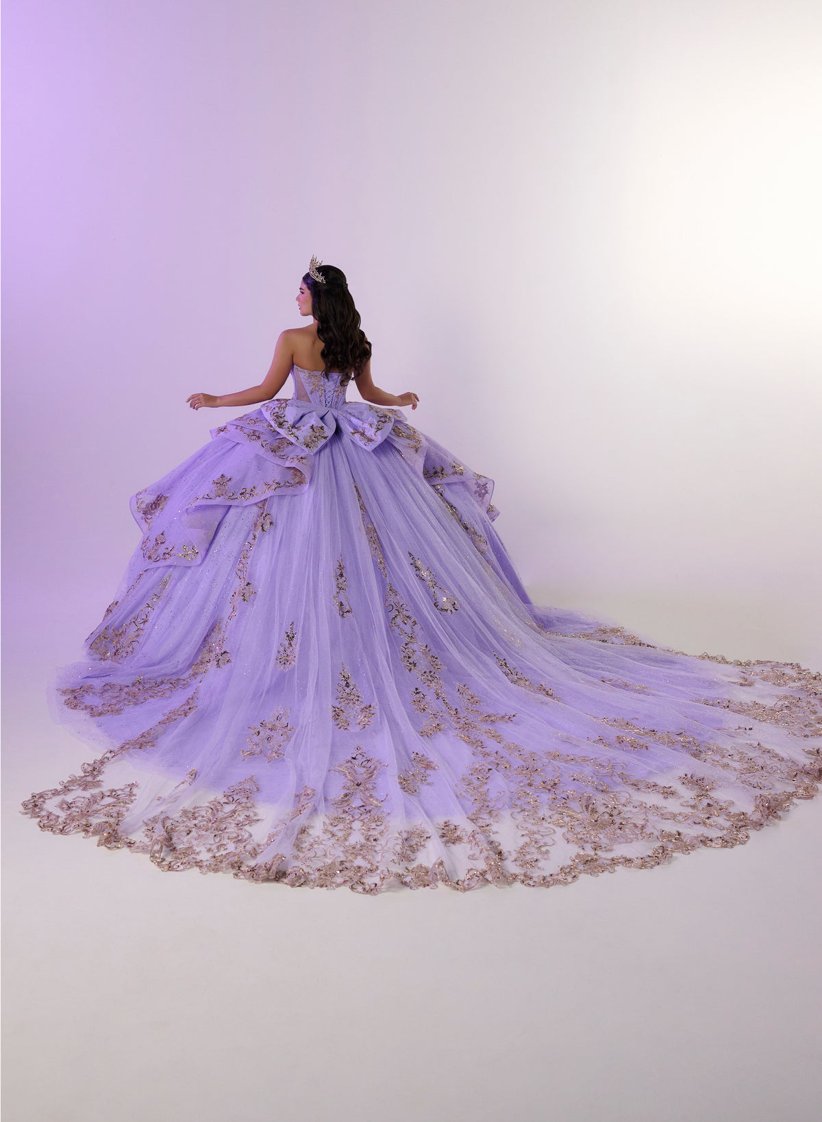 A model viewed from behind wearing a voluminous lavender quinceañera dress with gold floral embroidery and a long matching train, set against a soft purple studio backdrop. / Regal ball gown featuring intricate embroidered appliqués, a structured bodice, dramatic layered overskirt details, and a sweeping cathedral-length train for a royal-inspired look.