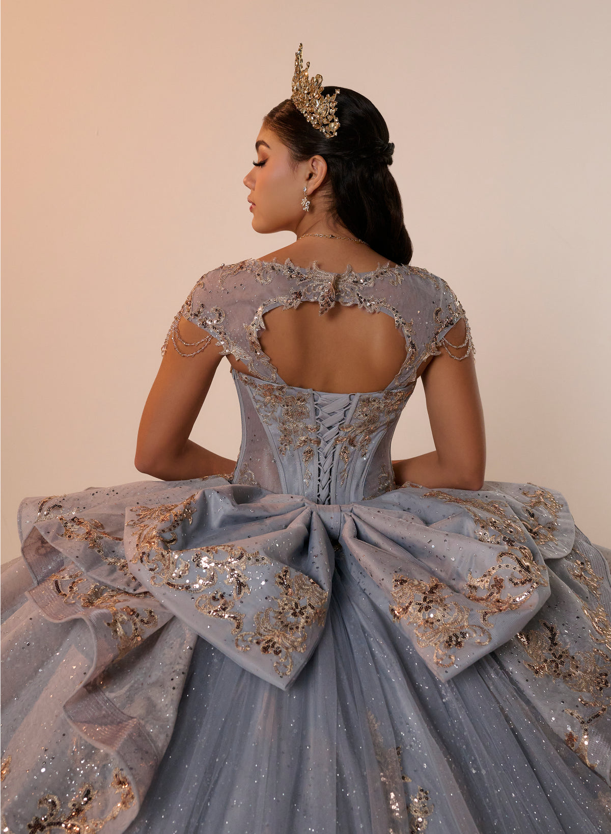 A woman photographed from the back wearing an elaborate light blue ball gown with gold sequin embellishments, a large bow at the back, and a gold tiara against a plain beige studio background. / Regal ball gown featuring intricate embroidered appliqués, a structured bodice, dramatic layered overskirt details, and a sweeping cathedral-length train for a royal-inspired look.