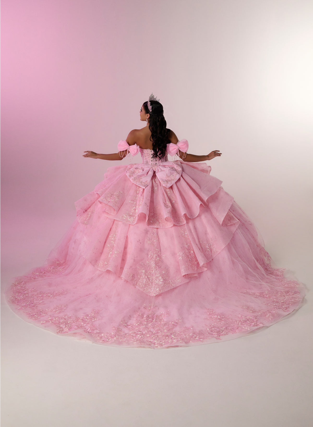 A woman viewed from behind wearing an elaborate off-the-shoulder pink ball gown with a large bow at the back and a full layered tulle skirt, posed against a soft pink-to-white studio backdrop. / Princess-inspired ball gown featuring a sweetheart corset bodice, intricate floral appliqués, dramatic layered overskirt details, and a voluminous skirt with a statement bow at the back.