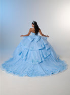 A woman wearing a voluminous light blue quinceañera ball gown, photographed from behind against a soft blue-to-white studio backdrop. / Princess-inspired ball gown featuring a sweetheart corset bodice, intricate floral appliqués, dramatic layered overskirt details, and a voluminous skirt with a statement bow at the back.
