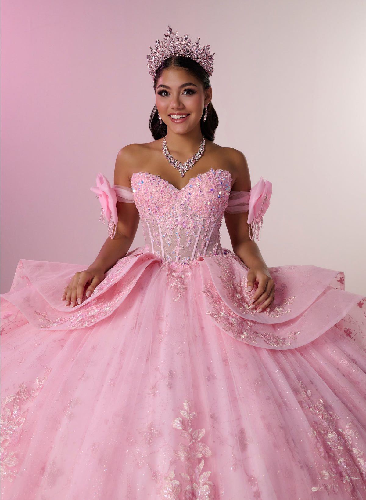 A young woman wearing a voluminous pink ball gown with off-the-shoulder straps, a jeweled corset bodice, and a sparkling tiara, posed against a soft pink studio background. / Princess-inspired ball gown featuring a sweetheart corset bodice, intricate floral appliqués, dramatic layered overskirt details, and a voluminous skirt with a statement bow at the back.