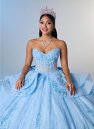 A woman wearing a strapless light blue quinceañera ball gown with intricate lace and sequin embellishments, a matching tiara, and a necklace, posing against a light gray studio background. / Princess-inspired ball gown featuring a sweetheart corset bodice, intricate floral appliqués, dramatic layered overskirt details, and a voluminous skirt with a statement bow at the back.