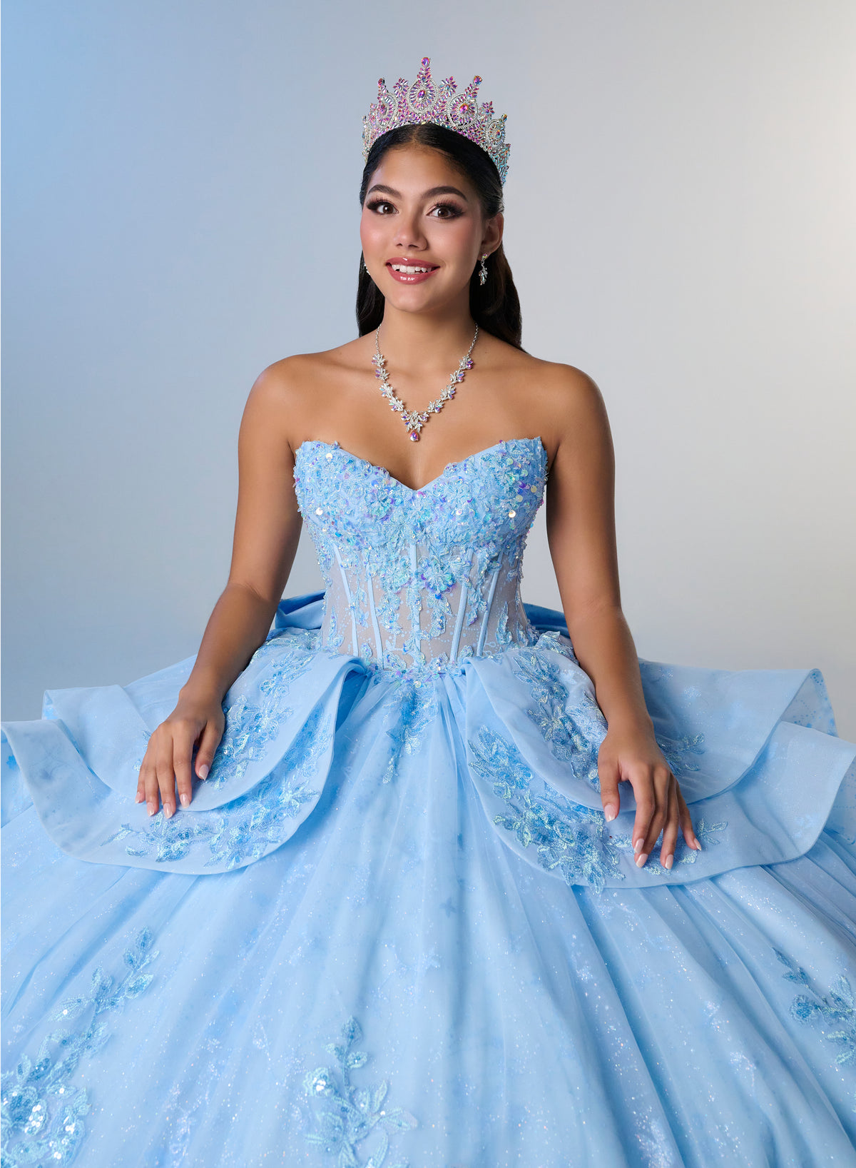 A woman wearing a strapless light blue quinceañera ball gown with intricate lace and sequin embellishments, a matching tiara, and a necklace, posing against a light gray studio background. / Princess-inspired ball gown featuring a sweetheart corset bodice, intricate floral appliqués, dramatic layered overskirt details, and a voluminous skirt with a statement bow at the back.