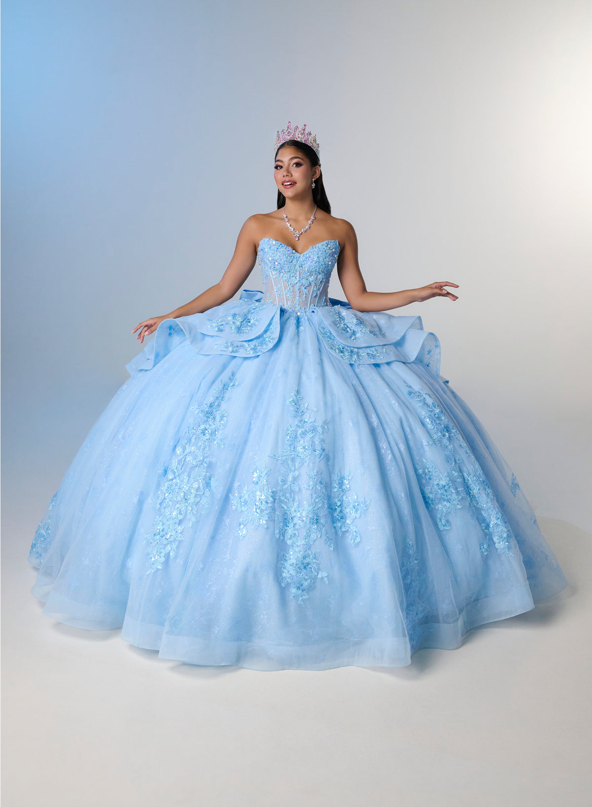 A woman wearing a voluminous light blue ball gown with floral lace appliqué and a sweetheart neckline, posed against a soft blue-to-white studio backdrop. She is wearing a small tiara and necklace. / Princess-inspired ball gown featuring a sweetheart corset bodice, intricate floral appliqués, dramatic layered overskirt details, and a voluminous skirt with a statement bow at the back.