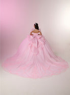 A young girl viewed from behind wearing a voluminous light pink ball gown with a large bow on the back, set against a soft pink and white studio backdrop. / Fairytale ball gown featuring a crystal-embellished corset bodice, elegant cape sleeves, dramatic layered skirt details, and a voluminous silhouette finished with a statement bow and sweeping train.