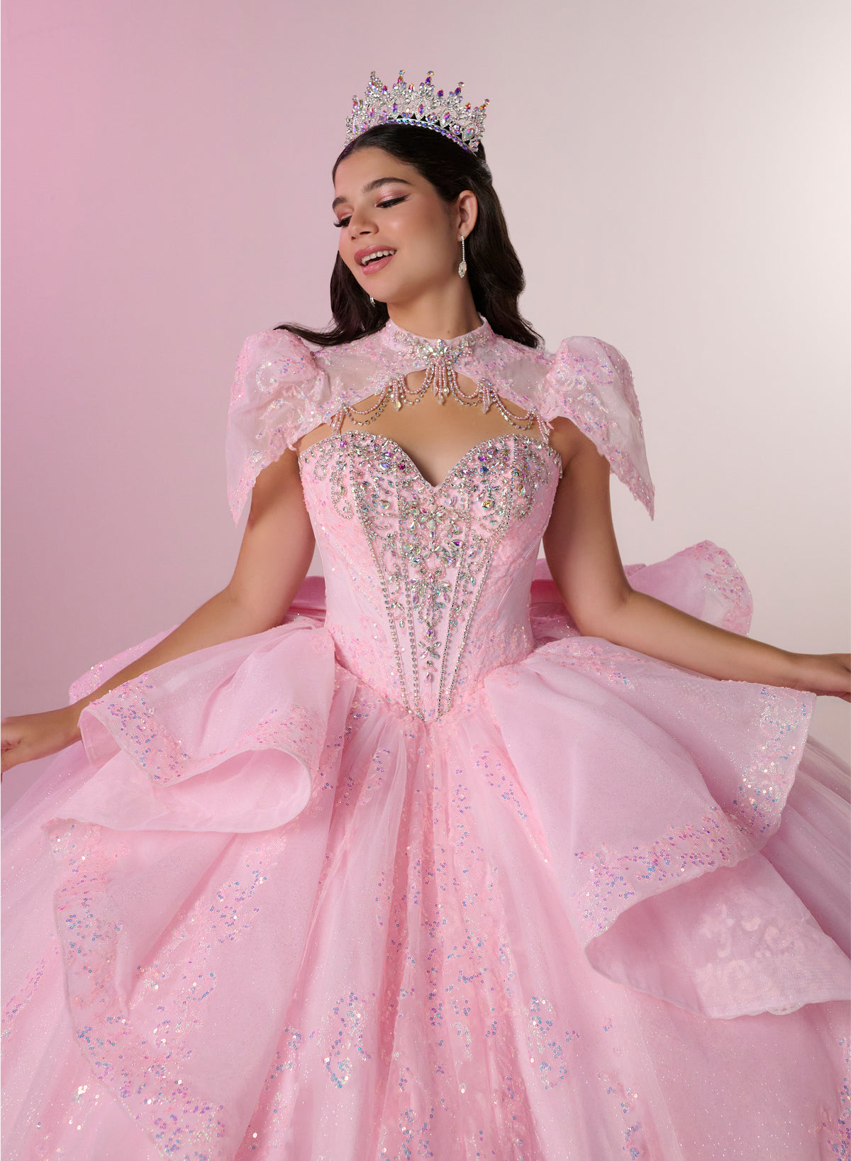 A young woman wearing a sparkling light pink ball gown with sheer puff sleeves, a beaded corset bodice, and a full layered skirt, posed against a soft pink studio background with a jeweled tiara. / Fairytale ball gown featuring a crystal-embellished corset bodice, elegant cape sleeves, dramatic layered skirt details, and a voluminous silhouette finished with a statement bow and sweeping train.