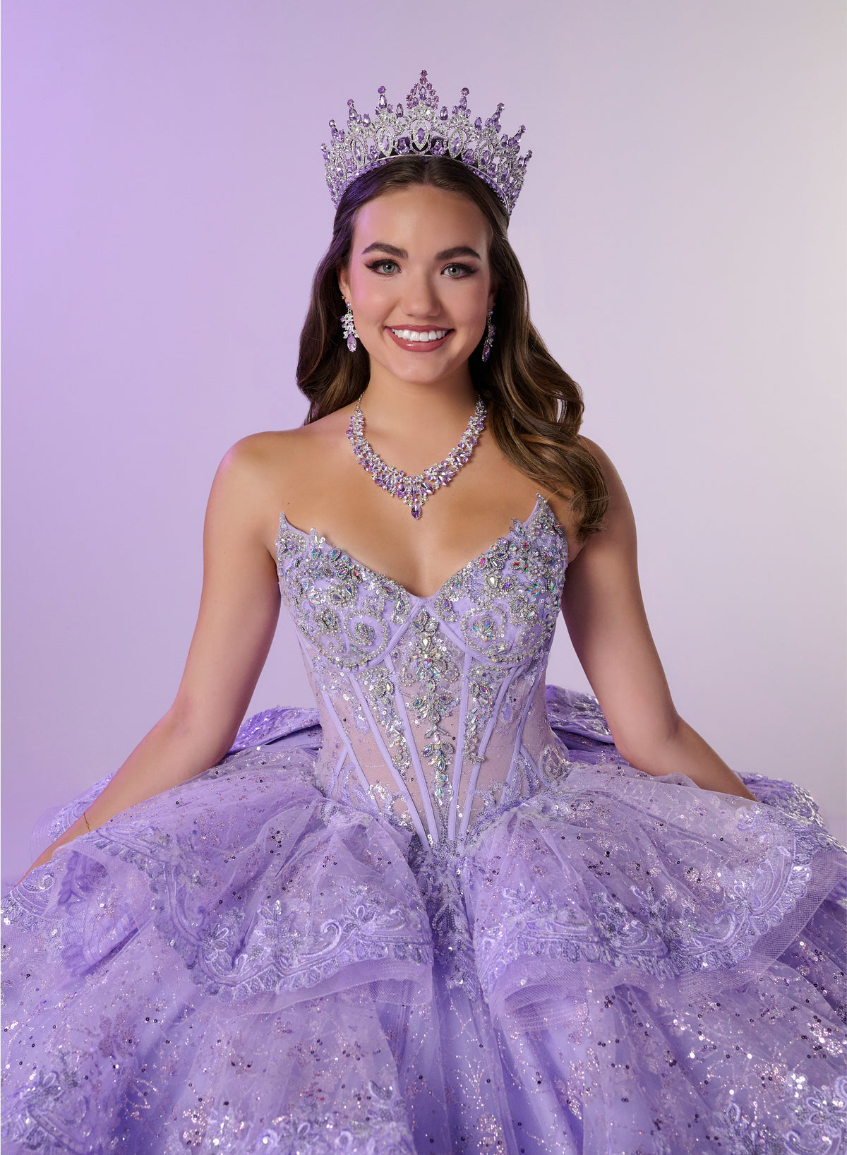 A smiling young woman wearing a sparkling lavender quinceañera-style gown with a fitted embellished bodice and voluminous tulle skirt, along with a jeweled tiara and matching necklace, posed against a soft lavender studio background. / Enchanting ball gown featuring a structured sweetheart corset bodice, shimmering embroidered details, dramatic layered ruffles, and a sweeping cathedral-length train for a regal princess-inspired look.
