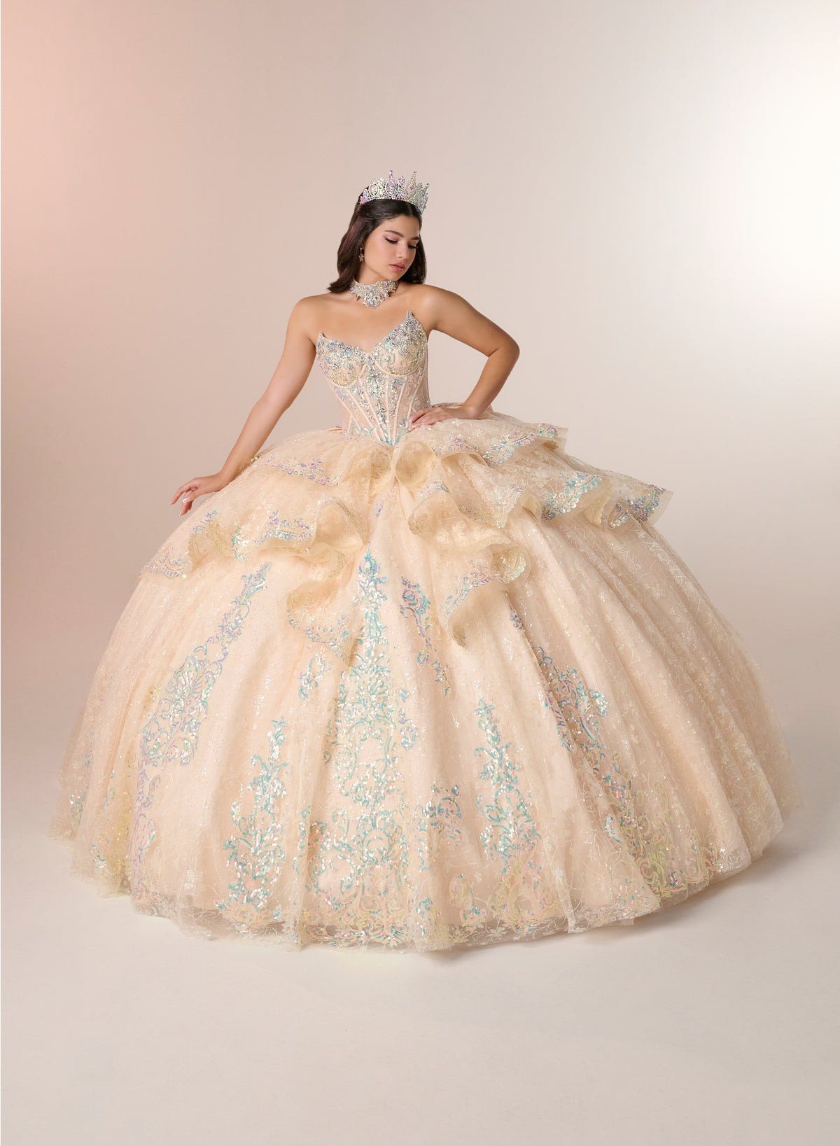 A woman wearing a voluminous champagne-beige ball gown with intricate silver and blue floral embroidery, posing in a studio setting with a soft beige background. / Enchanting ball gown featuring a structured sweetheart corset bodice, shimmering embroidered details, dramatic layered ruffles, and a sweeping cathedral-length train for a regal princess-inspired look.