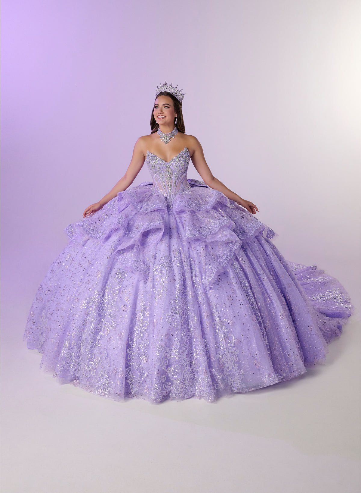 A woman wearing a voluminous lavender quinceañera ball gown with silver glitter and lace detailing, a matching tiara, and a necklace, posing against a light lavender studio backdrop. / Enchanting ball gown featuring a structured sweetheart corset bodice, shimmering embroidered details, dramatic layered ruffles, and a sweeping cathedral-length train for a regal princess-inspired look.