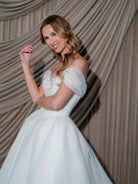 A woman wearing an off-the-shoulder white wedding gown with a voluminous sparkle tulle skirt, posing in front of draped beige fabric. / Ball gown in two face organza with glitzy underlayer, sweetheart neckline and off shoulder straps. Lace up back closure.