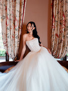 A woman wearing a strapless white wedding gown with a structured lace bodice and voluminous tulle skirt, posed indoors in front of patterned curtains and a brown wall. / Semi sheer basque waist bodice with lace appliques and dramatic tulle skirt.