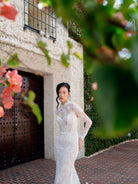 A woman wearing a fitted white lace wedding gown stands outdoors in front of a stucco building with a dark wooden door, framed by green leaves and pink flowers in the foreground. / All over lace and lace applique slim gown with detachable lining and jacket as well as structured corset.
