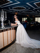 A woman in a dramatic off-the-shoulder white satin gown with a long train leans against a bar counter in an elegant, dimly lit venue with a dark patterned ceiling and a white candelabra in the foreground. / Off the shoulder sculpted neckline gown with basque waist and full, gathered skirt.
