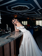 A woman in a white off-the-shoulder gown leans on a marble bar counter in a dimly lit venue with a dark geometric ceiling. / Off the shoulder sculpted neckline gown with basque waist and full, gathered skirt.