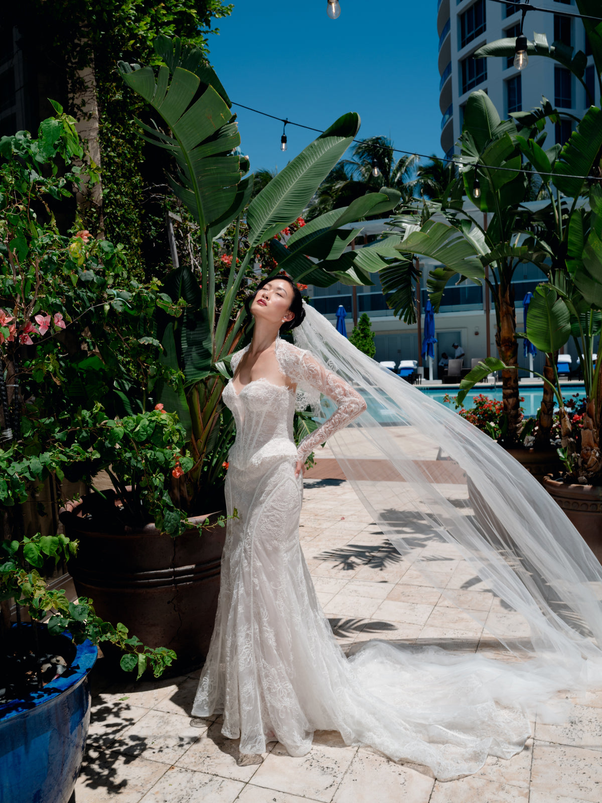 A woman wearing a white lace wedding gown with a long sheer veil stands outdoors on a tiled walkway beside a pool, surrounded by tropical plants and blue sky. / Slim lace gown with semi-sheer bodice with sweetheart neckline and bustier cut. Small peplums on sides hips. Invisible zipper back closure. Matching lace bolero with long sleeves.