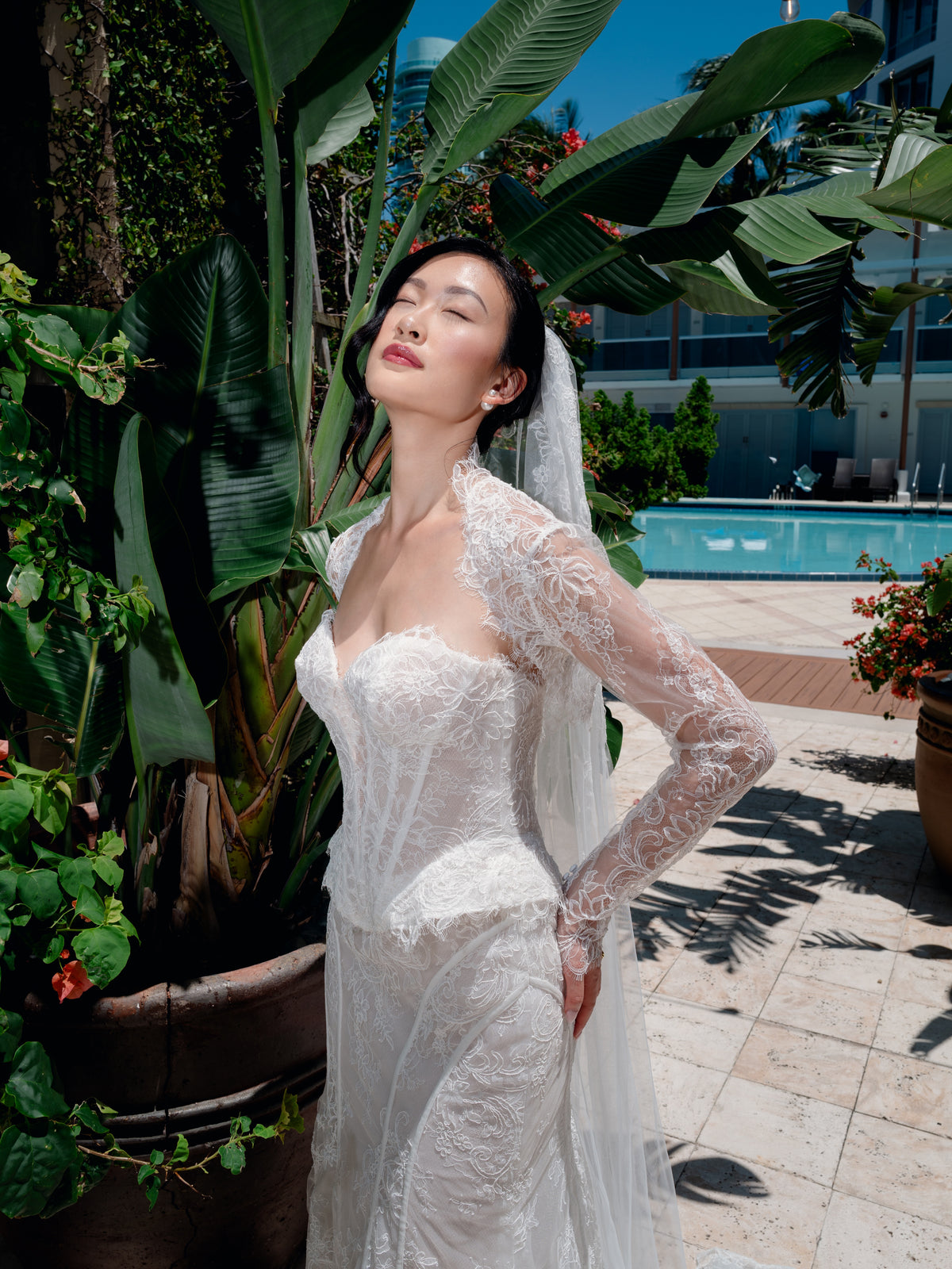 A woman wearing a white lace bridal gown with sheer long sleeves and a veil, standing outdoors beside a tropical pool with large green plants and tiled decking. / Slim lace gown with semi-sheer bodice with sweetheart neckline and bustier cut. Small peplums on sides hips. Invisible zipper back closure. Matching lace bolero with long sleeves.