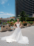 A woman wearing a strapless white wedding gown with a long sheer overskirt and train, standing outdoors on a tiled walkway with a resort pool, palm trees, and a tall hotel building in the background. / Satin mermaid gown with lace appliques and detachable overskirt.