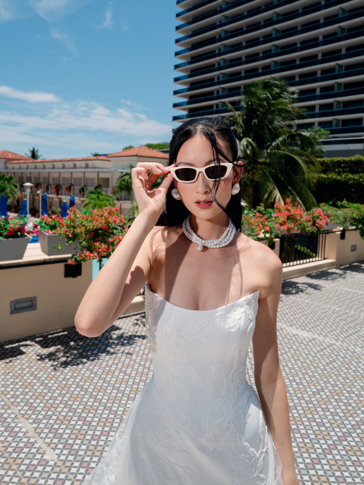 A woman wearing a white strapless dress and white cat-eye sunglasses stands outdoors on a tiled patio with palm trees and a modern high-rise building in the background. / Satin mermaid gown with lace appliques and detachable overskirt.