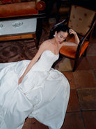 A woman reclining on a terracotta tile floor in a white strapless satin wedding gown, with an orange upholstered chair and a wooden table in the background. / Dramatic drop waist, double face satin ballgown with placed lace and lace up back detail.