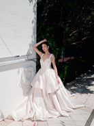 A woman wearing a voluminous ivory satin wedding gown with a sweetheart neckline and layered skirt, posing outdoors against a white stucco wall and green foliage. / Dramatic, sculpted mikado gown with lace placement.
