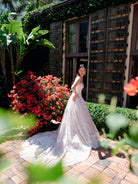 A woman wearing a sleeveless white lace wedding gown with a long train stands outdoors on a brick patio beside a rustic wooden building, with red flowers and green foliage in the background. / All over multicolored lace A-line gown.