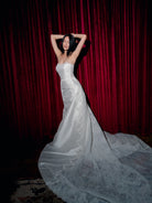A woman wearing a strapless white wedding gown with a fitted bodice, sheer lace overlay, and a long train, posing with her hands behind her head in front of a dark red velvet curtain. / Slim mikado style with pleated bodice with straight neckline and zipper/buttons back closure. Lace scallop decoration on bodice and on skirt sides. Matching mikado detachable train with sheer back area covered with lace.