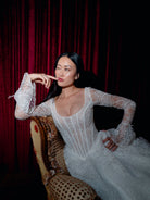 A woman wearing a white lace long-sleeve gown sits on an ornate upholstered chair in front of a dark red velvet curtain. / All over lace gown with long sleeve, basque waist and A-line skirt with lace up back.
