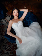 A woman reclining on a dark blue velvet armchair wearing a strapless white lace wedding gown in a dimly lit indoor setting. / Organza and lace princess ballgown with lace applique details.