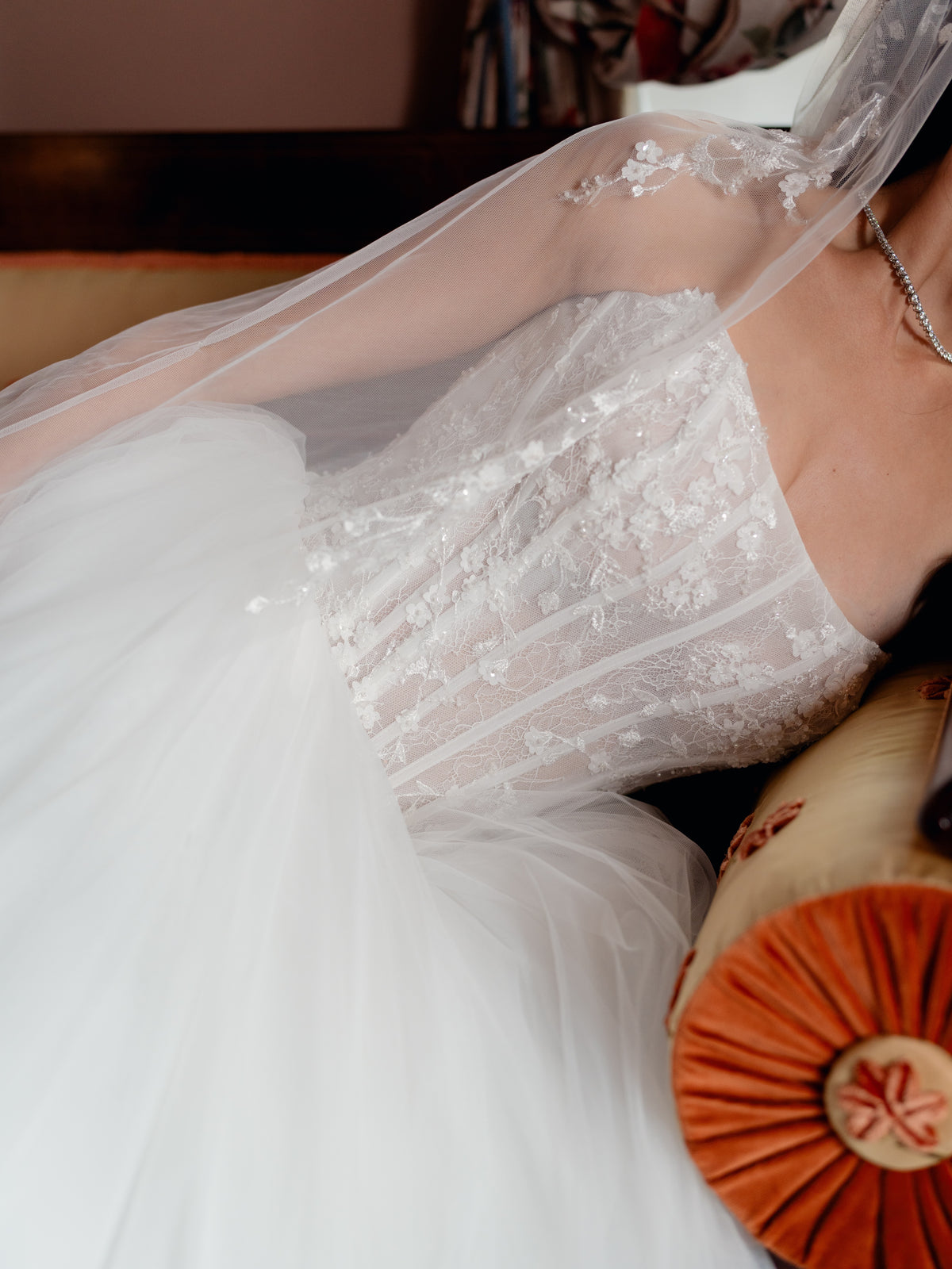 A close-up of a white bridal gown with floral lace applique and a sheer veil, worn by a bride seated indoors next to a rust-colored pillow. / Semi sheer basque waist bodice with lace appliques and dramatic tulle skirt.