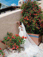 A woman in a white strapless wedding gown with a long sheer veil poses outdoors against a beige stucco wall, surrounded by large planters filled with red flowers.
