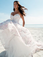 A woman wearing a strapless white wedding gown with pink floral embroidery and a full tulle skirt, standing on a sandy beach with the ocean and blue sky in the background. / All over multicolored lace A-line gown.