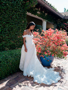 A woman wearing an off-the-shoulder white lace wedding gown stands on a brick patio beside a large blue planter filled with pink flowers, with a leafy wall and house in the background. / All over lace column gown with a dramatic matching overskirt.