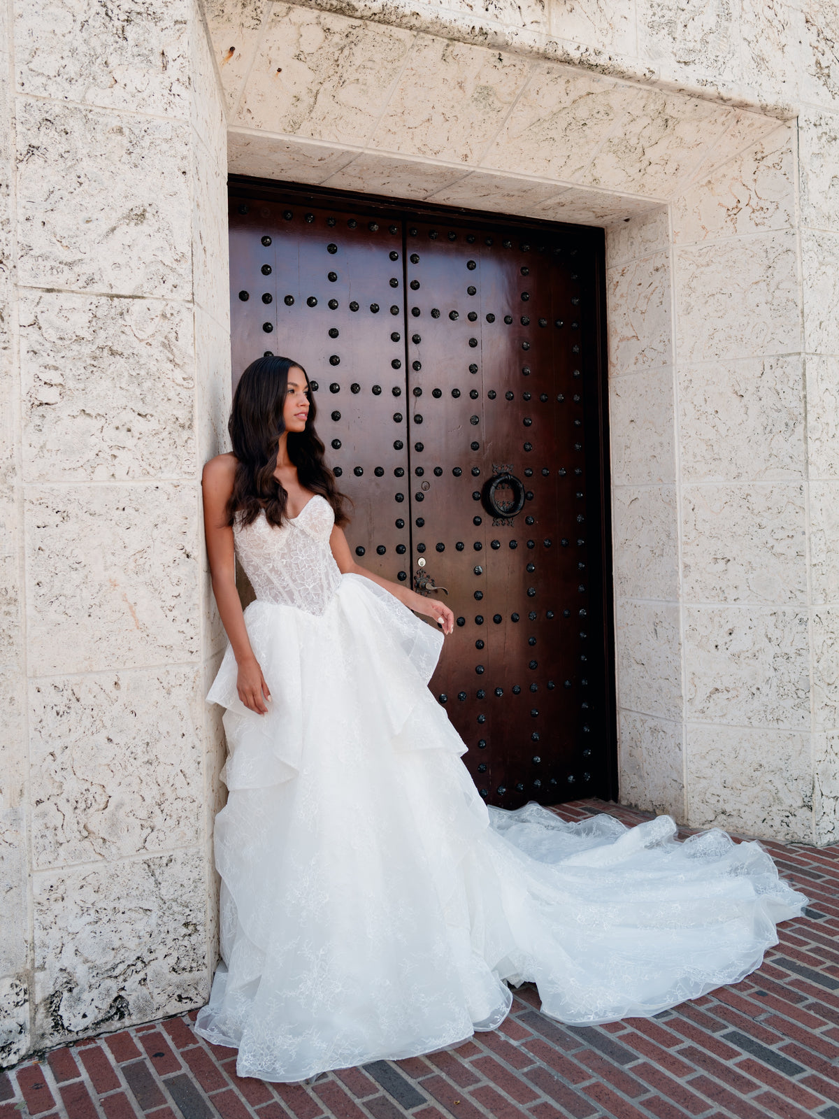 A woman wearing a strapless white wedding gown with a lace bodice and full layered tulle skirt stands beside a large dark wooden door set in a stone building. / All over lace gown with cat eye neckline and soft, dramatic cascading skirt.