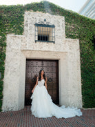 A woman wearing a white strapless wedding gown with a long train stands in front of a stone doorway with ivy-covered walls and dark wooden doors. / All over lace gown with cat eye neckline and soft, dramatic cascading skirt.