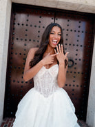 A woman in a white lace wedding gown stands in front of a large dark wooden door, pointing to a ring on her finger with a joyful expression. / All over lace gown with cat eye neckline and soft, dramatic cascading skirt.